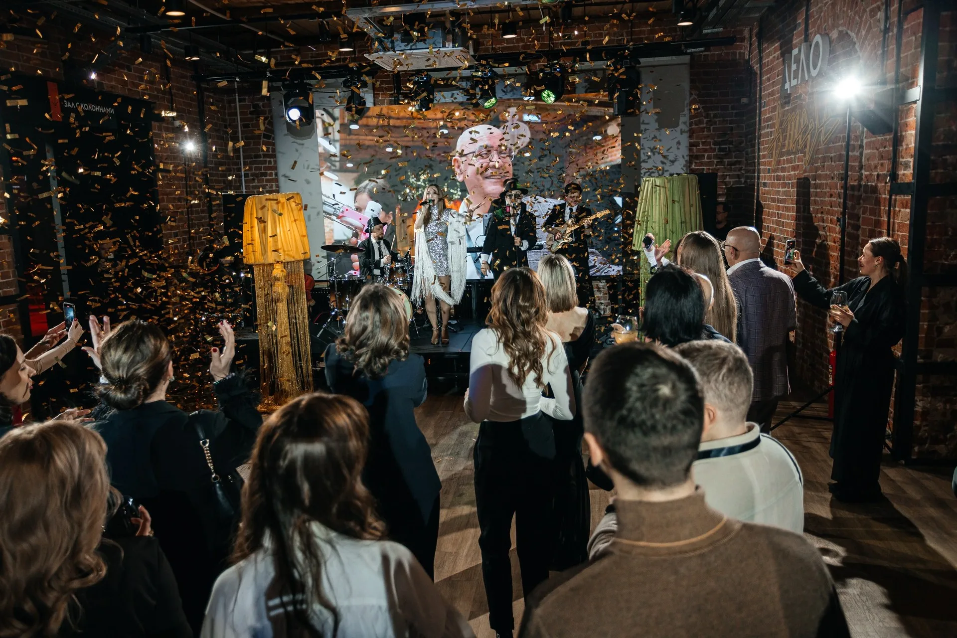 Две женщины позируют на фоне золотого текстильного шатра с длинной бахромой; вокруг — хвойные ветви и подсвеченные гирляндами деревья.