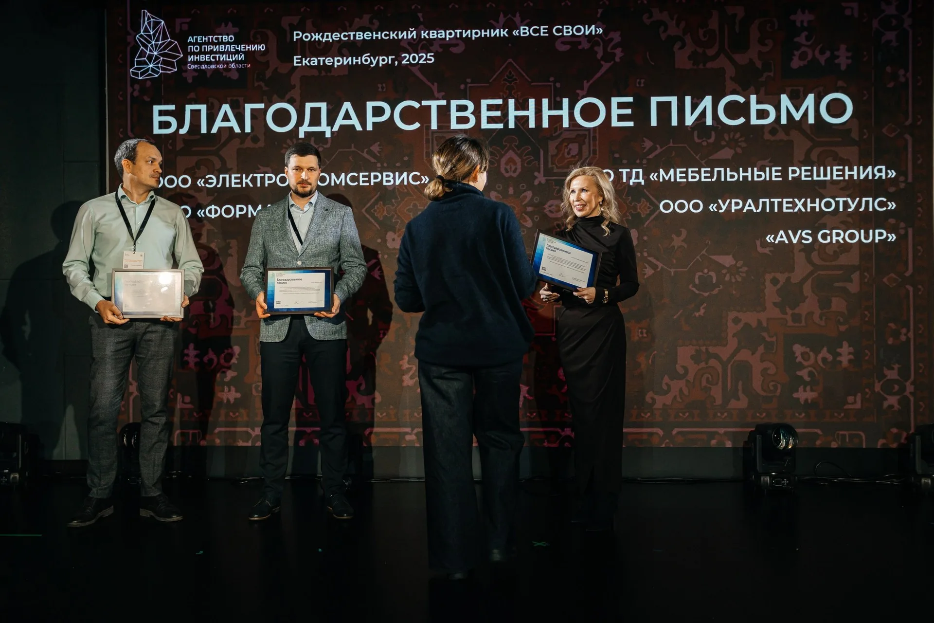 Две женщины позируют на фоне золотого текстильного шатра с длинной бахромой; вокруг — хвойные ветви и подсвеченные гирляндами деревья.