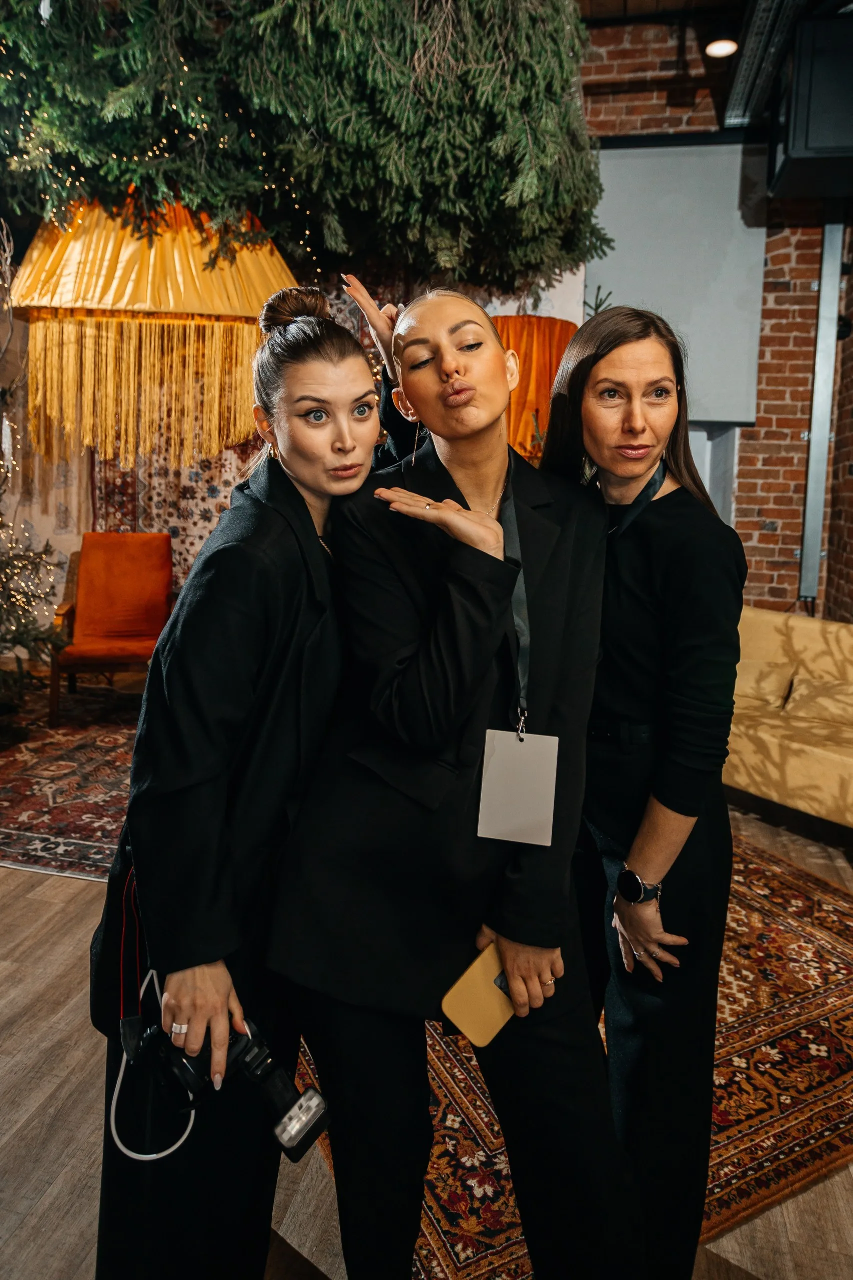 Две женщины позируют на фоне золотого текстильного шатра с длинной бахромой; вокруг — хвойные ветви и подсвеченные гирляндами деревья.