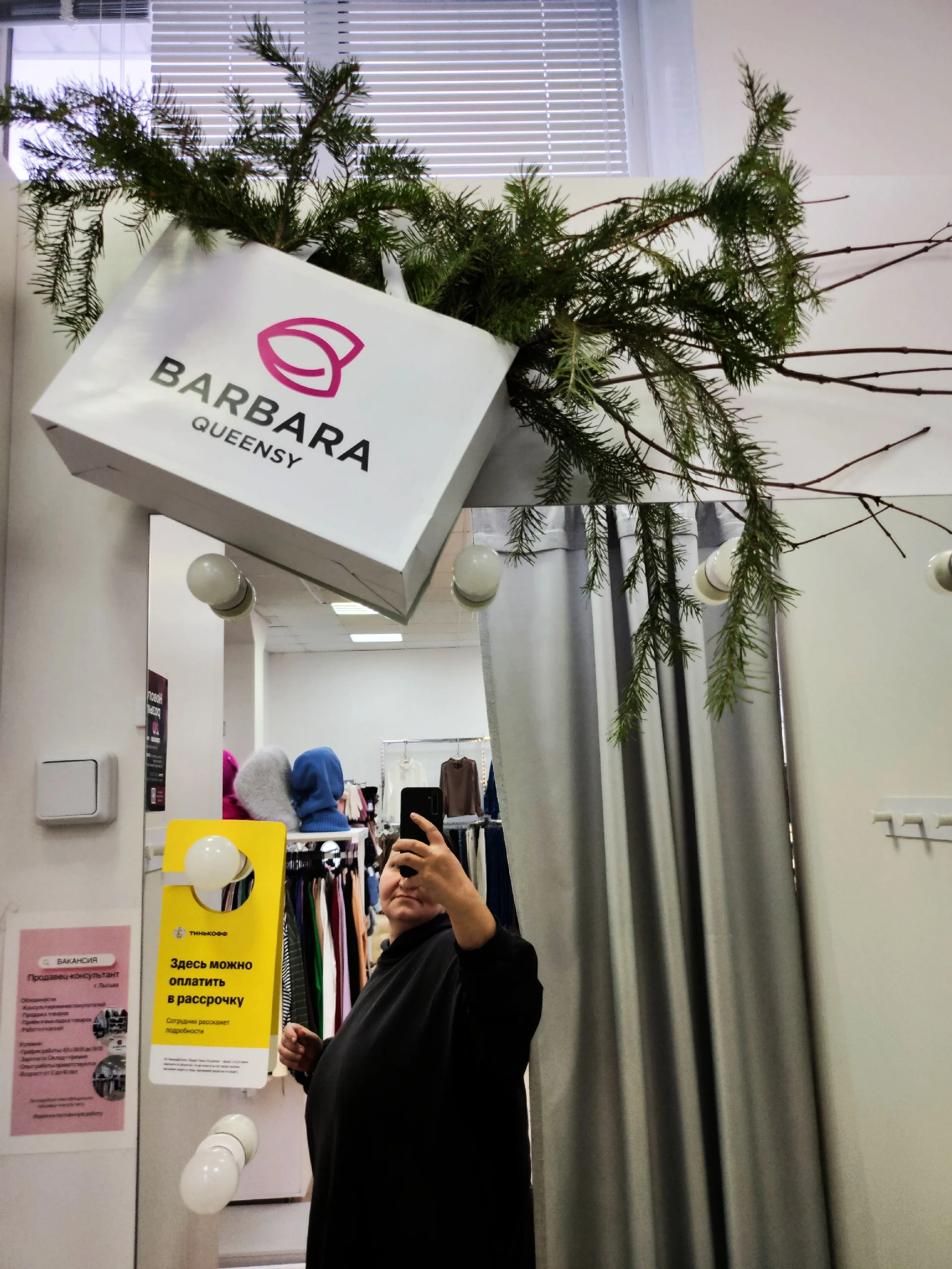 Пышная новогодняя композиция из сосновых ветвей с тёплой гирляндой и ярко-розовыми и чёрными шарами возле серого дивана.